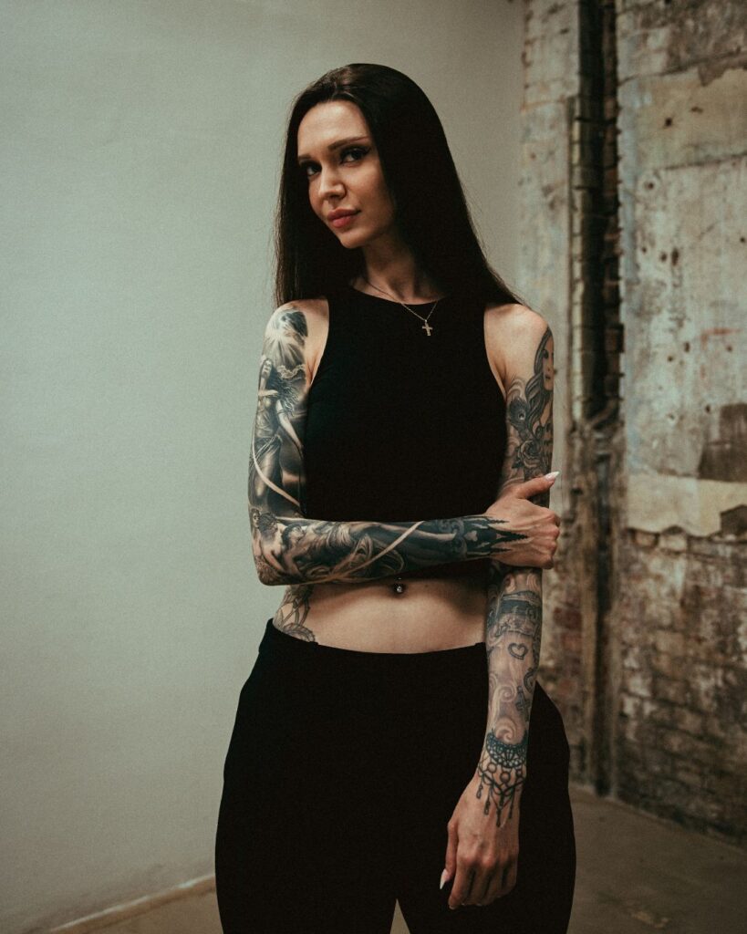 A woman with long dark hair stands in front of a white brick background, wearing black, she has two tattoo sleeves, one arm is holding the other .