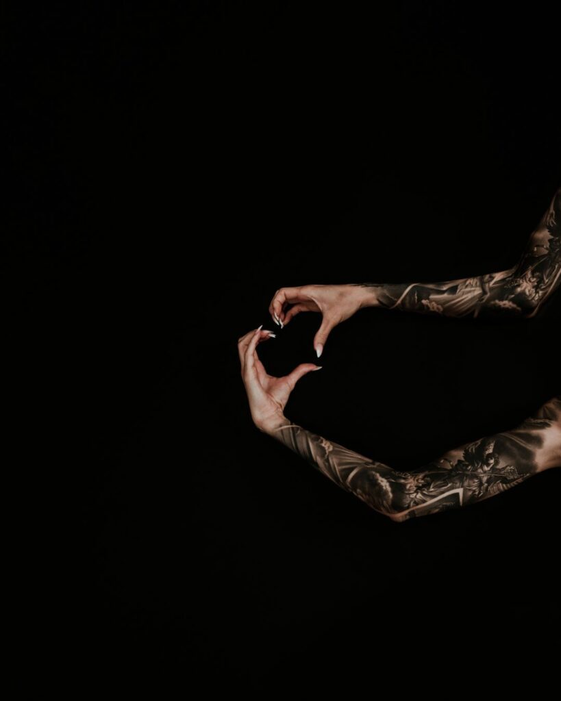 Two arms on a black background, both with black and grey realism religious tattoos. The hands are creating the shape of a heart.