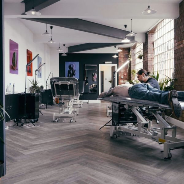 No Regrets Birmingham tattoo studio: The image shows a long room with wooden flooring and lots of natural light. At the forefront you can see a tattoo artist tattooing a client.