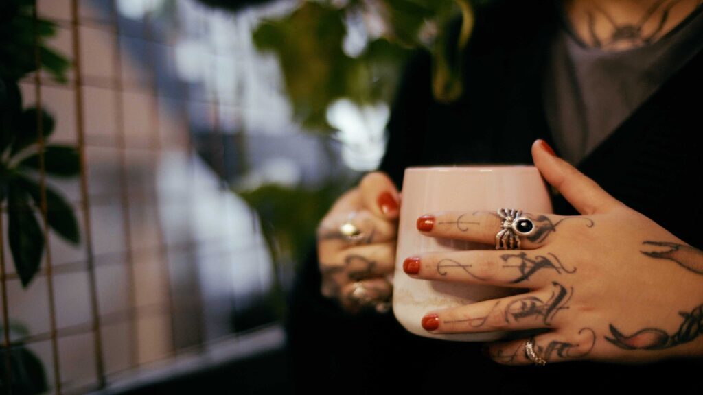 Woman with tattooed hands and red nails holding a pink coffee mug.