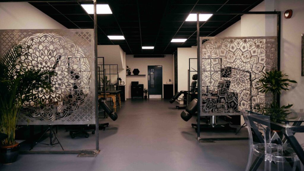 Landscape image of No Regrets Worcester. It shows two metal room dividers that has a cut out mandala design on. In the background you can see three tattoo chairs, a back wall with a door in the middle and small cabinets. 