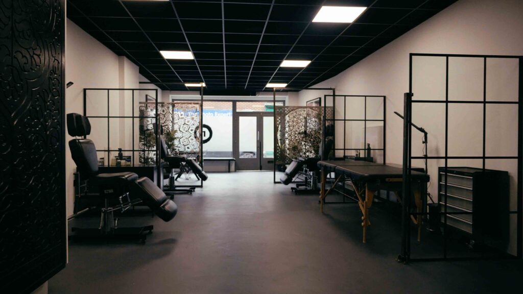 Image shows studio from the back of the studio. It has glass and metal square room dividers, three tattoo chairs and a tattoo bed. 