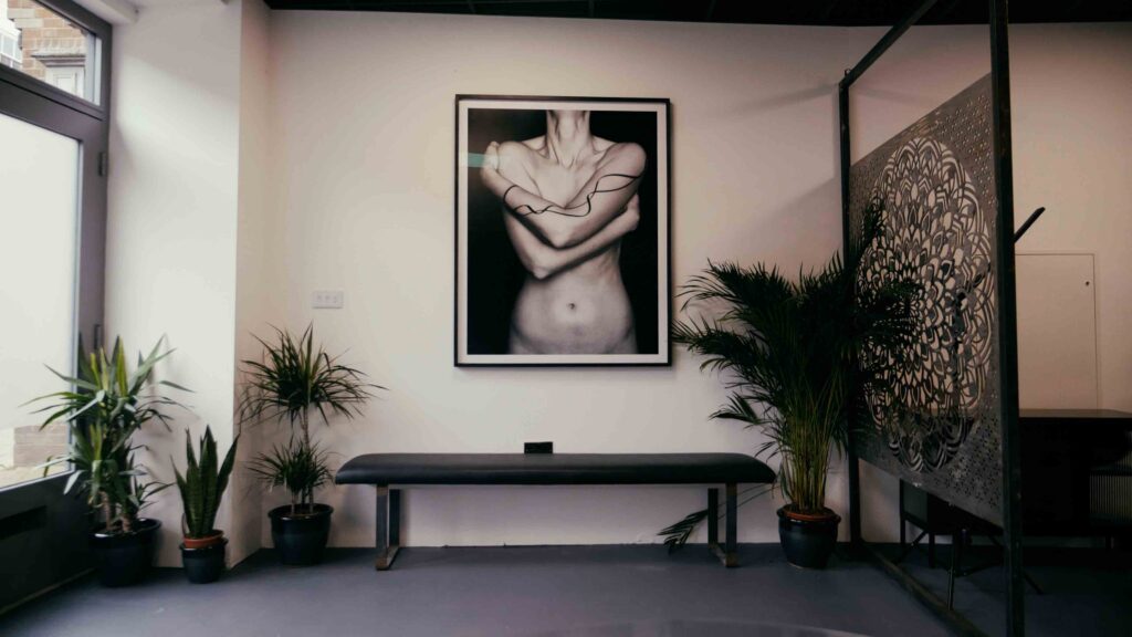 Image shows reception of Worcester tattoo studio. A long bench is in the middle of the frame with a black and white photograph image of a woman's torso, arms covering breasts. Four green plans are situated around the plant. 