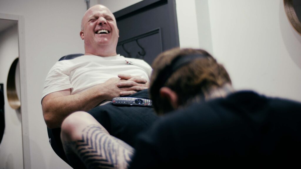 Joe sits in the tattoo chair at No Regrets Cheltenham laughing while Ash Grey tattoos his foot