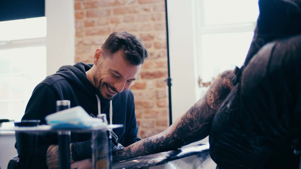 male tattoo artist smiling while tattooing black and grey arm sleeve of client.