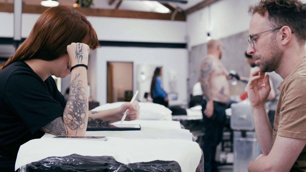 female tattoo artist drawing design on Ipad while client sits opposite watching.