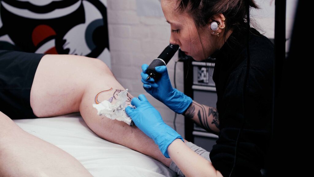 A tattoo artist with earphones and blue gloves is using a tattoo machine to design a colorful tattoo on a client's leg. The client, mindful of what to wear when getting tattooed is wearing shorts, allowing for a comfortable tattooing experience.