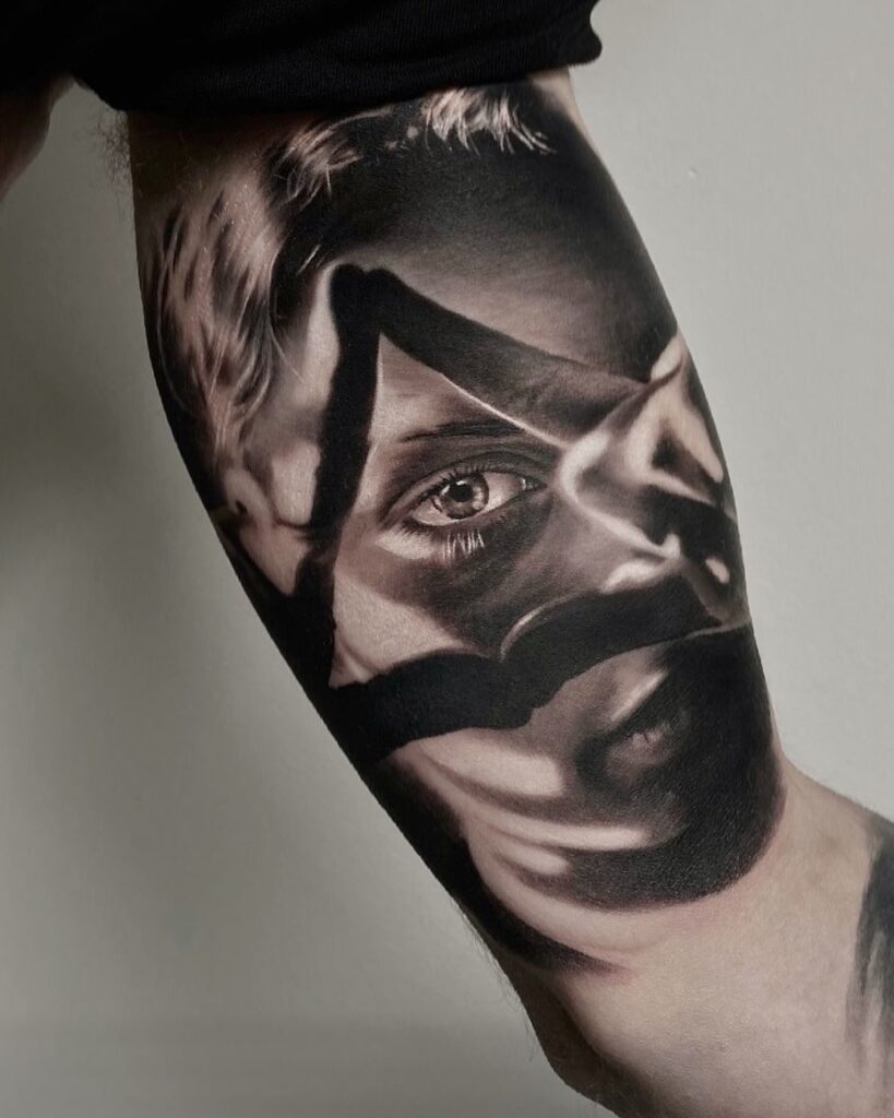 Tattooed arm against grey background. Tattoo shows realistic portrait of a woman looking through a triangle made from fingers joining together. 