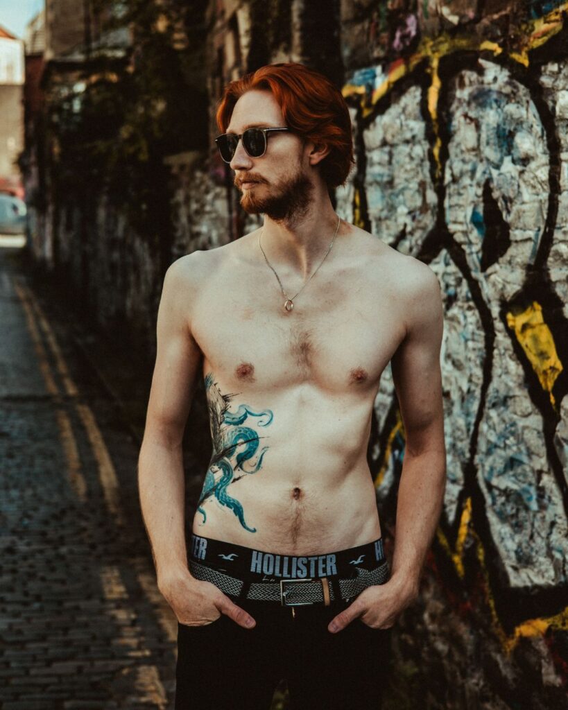 Joe stands in front of a brick wall which has graffiti on it. He is standing face on but looking to the right while wearing sunglasses. You can see his Kracken tattoo spanning his right side.