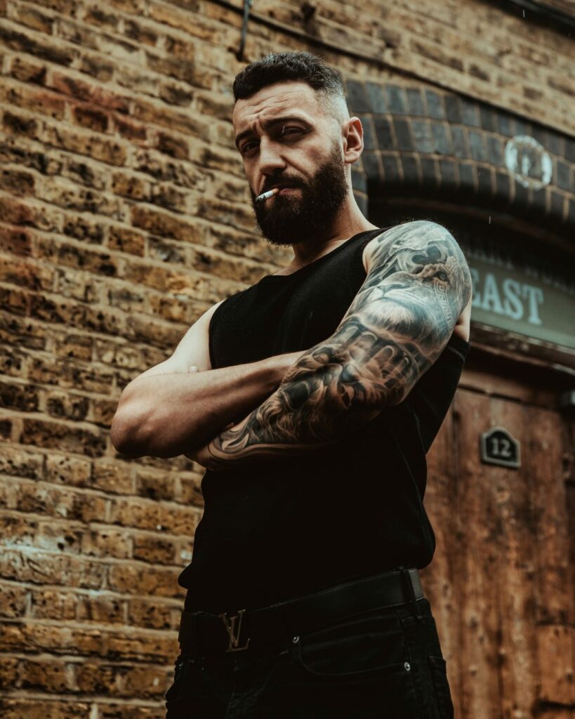 Adam stands with his arms crossed in front of a brick wall. He has a cigarette in his mouth and is looking down at the camera. You can see his black and grey tattoo sleeve on his left arm.