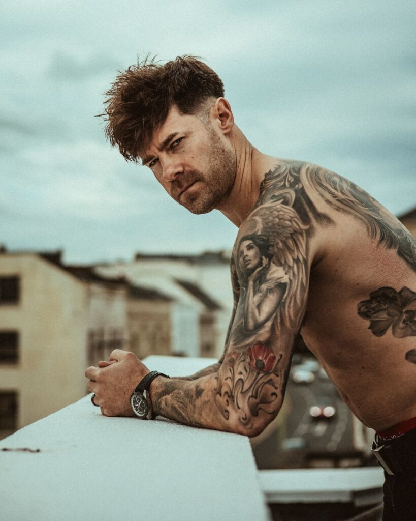 Rhys leans over a wall while looking into the camera from the right. His black and grey sleeve is in shot.