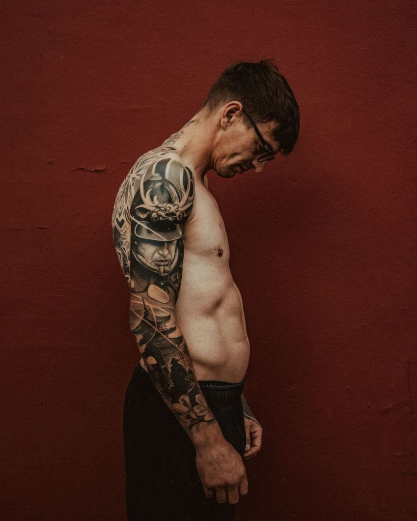 Dean stands side onto the camera while looking down. His right arm shows a black and grey tattoo sleeve of a Samurai and Tera temple.