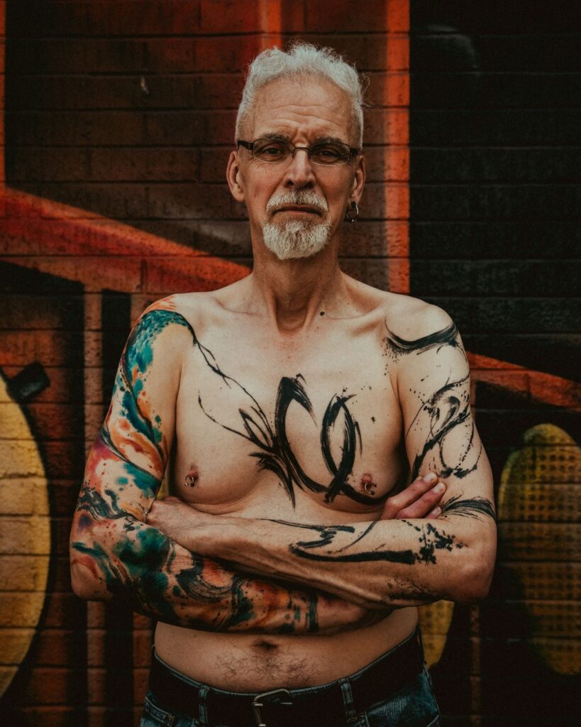 Steve, a slim, white man with white hair and glasses stands against a graffitied wall, topless with his arms crossed. His first tattoo - bold abstract paint strokes are shown going across both arms and chest