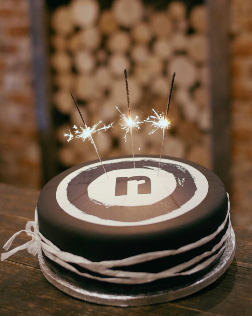 In the foreground we see a black and white birthday cake with the No Regrets logo with three lit sparklers on top.