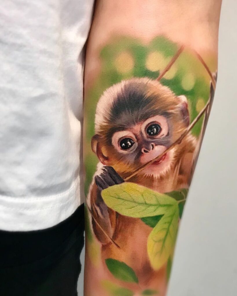 Colour realism forearm tattoo. Shows a baby monkey chewing on a branch with a green leafy background