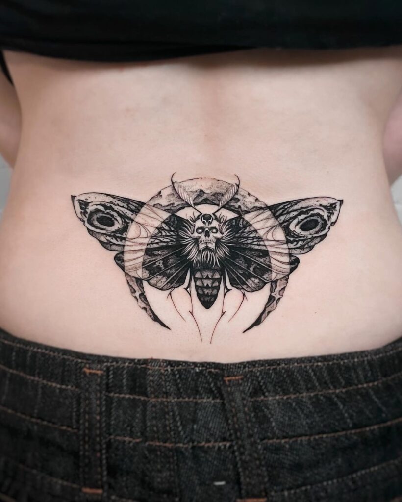 Lower back black and grey tattoos. Woman with lower back tattoo of a dark death moth with an upside down crescent moon in front of it. 